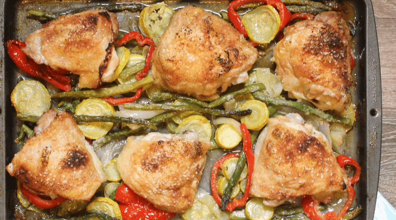 A sheet pan with six roasted chicken thighs laying on a bed of yellow squash and asparagus. 