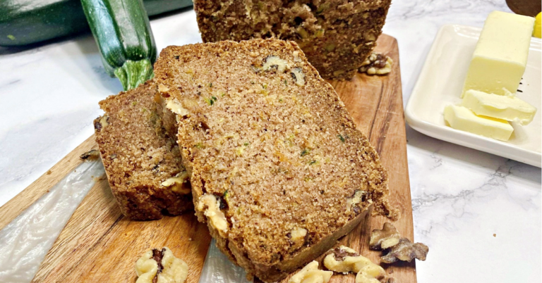 A image of two slices of zucchini bread stack on each other with the loaf behind them.